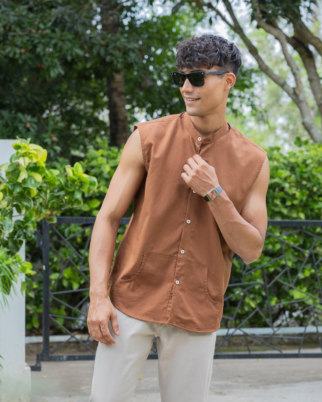 A man with short, curly hair wearing sunglasses, a sleeveless brown button-up Brunn - Sleeveless Shirt, and beige pants stands outdoors. He is looking down with his hands in his pockets. A backdrop of lush green foliage and trees is visible, showcasing his effortless style.