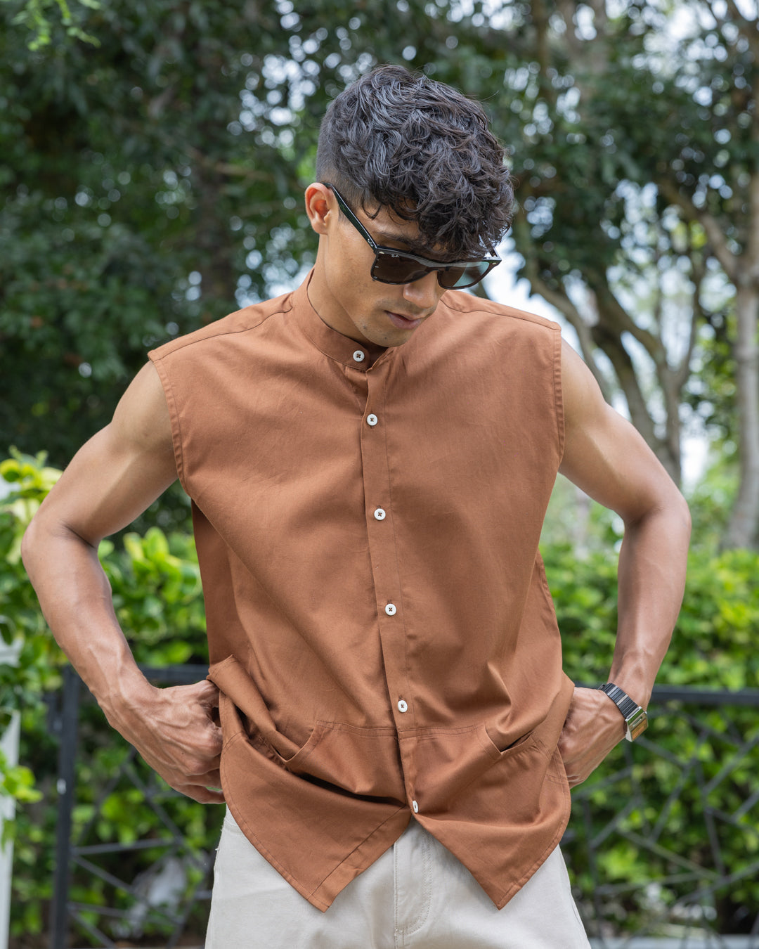 A man with short, curly hair wearing sunglasses, a sleeveless brown button-up Brunn - Sleeveless Shirt, and beige pants stands outdoors. He is looking down with his hands in his pockets. A backdrop of lush green foliage and trees is visible, showcasing his effortless style.