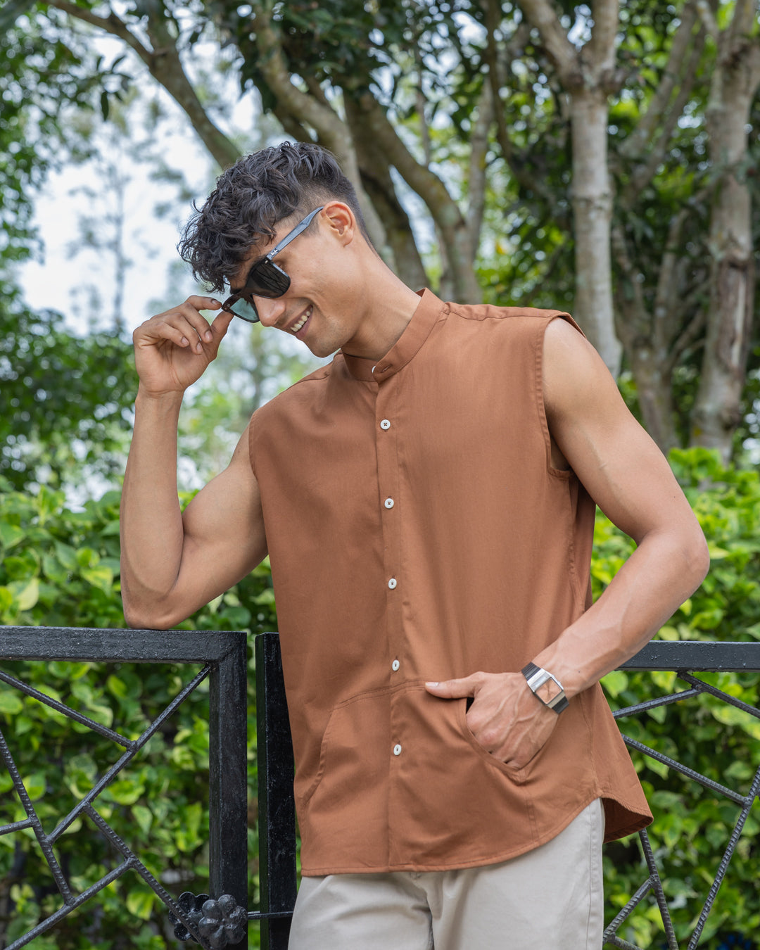 A man with short, curly hair wearing sunglasses, a sleeveless brown button-up Brunn - Sleeveless Shirt, and beige pants stands outdoors. He is looking down with his hands in his pockets. A backdrop of lush green foliage and trees is visible, showcasing his effortless style.