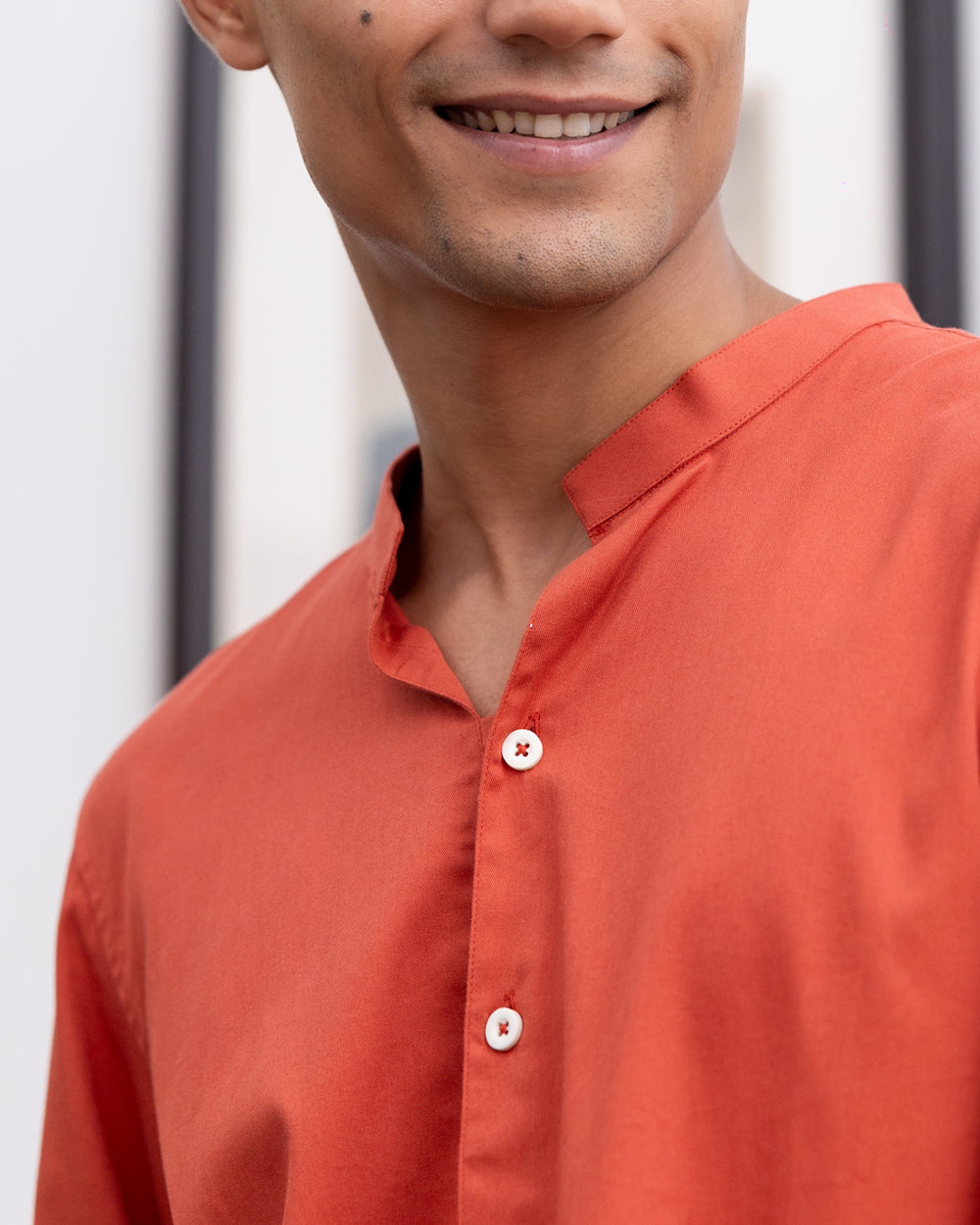 A young man with curly hair is standing indoors, wearing a Scarlatto - Short Kurta and beige pants. He is looking to his right with his left hand in his pocket. Behind him are a teal armchair, a framed picture on the wall, and a bookshelf with several books.