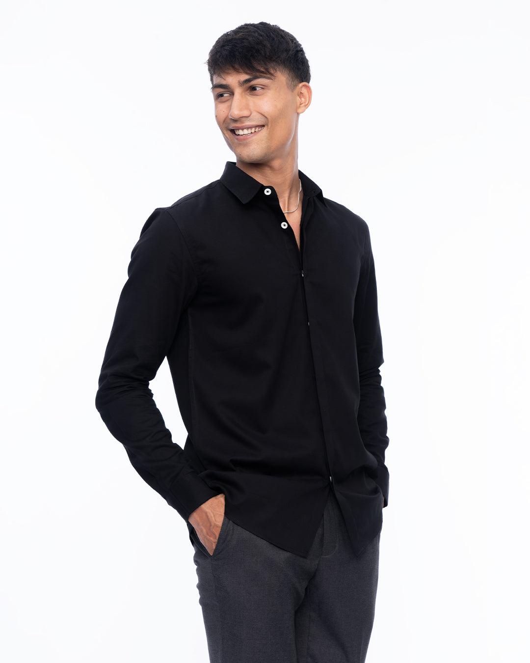 A person with short dark hair wearing an Obsidian - Classic Shirt made from premium cotton and gray pants is smiling while standing with their right hand in their pocket against a plain white background.