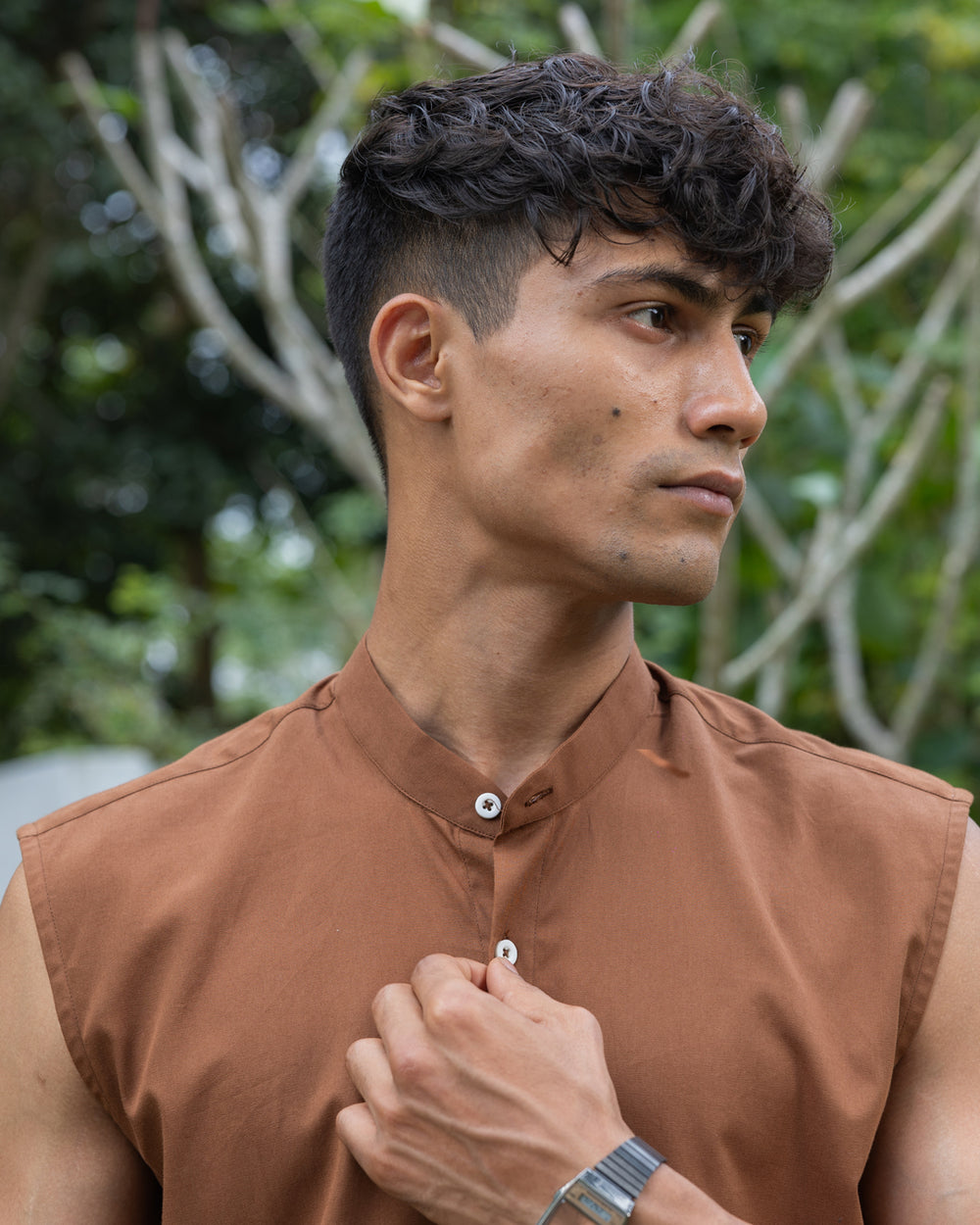 A man with short, curly hair wearing sunglasses, a sleeveless brown button-up Brunn - Sleeveless Shirt, and beige pants stands outdoors. He is looking down with his hands in his pockets. A backdrop of lush green foliage and trees is visible, showcasing his effortless style.