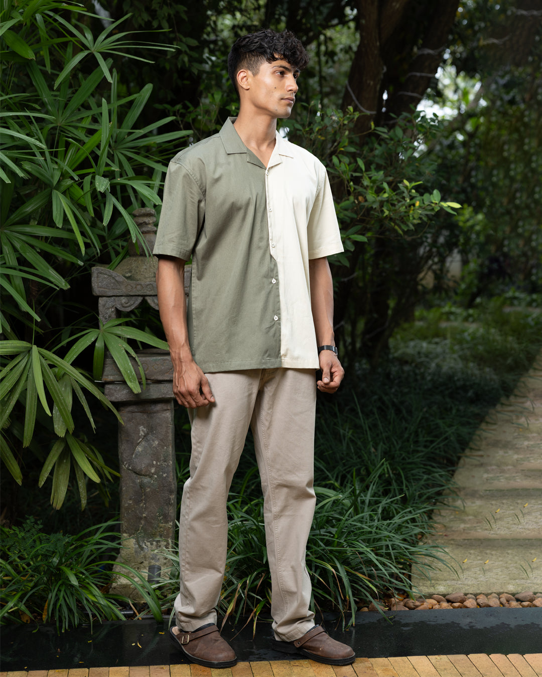 A man with curly dark hair smiles while standing in front of greenery. He is wearing a short-sleeved Dark Sage Moitie - Cuban Collar Shirt that is olive green on one side and cream on the other, paired with light-colored pants. He stands with one hand in his pocket.