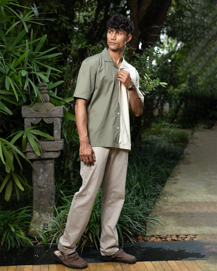 A man with curly dark hair smiles while standing in front of greenery. He is wearing a short-sleeved Dark Sage Moitie - Cuban Collar Shirt that is olive green on one side and cream on the other, paired with light-colored pants. He stands with one hand in his pocket.