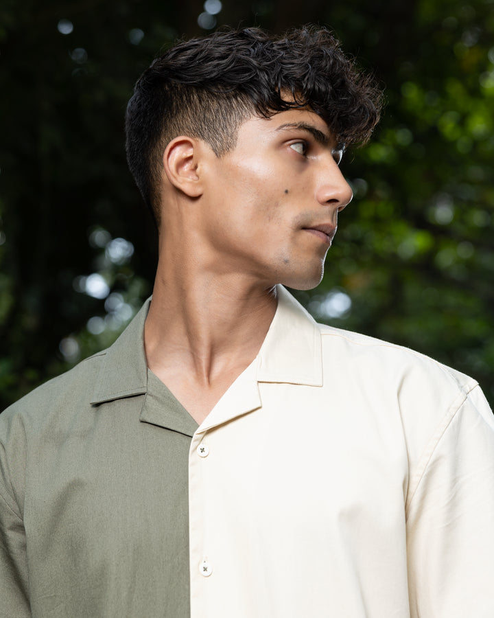 A man with curly dark hair smiles while standing in front of greenery. He is wearing a short-sleeved Dark Sage Moitie - Cuban Collar Shirt that is olive green on one side and cream on the other, paired with light-colored pants. He stands with one hand in his pocket.