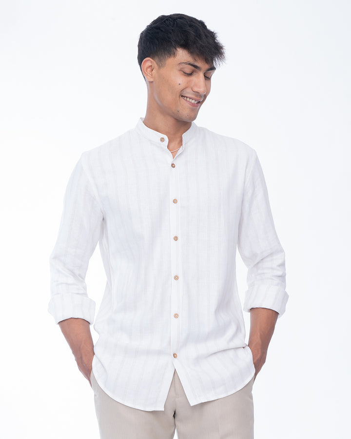 A person with short dark hair is smiling and wearing a Seashell White - Mandarin Shirt made from premium cotton. They have one hand in their pocket and the other hand is holding the edge of their shirt. The background is a plain white studio setting.