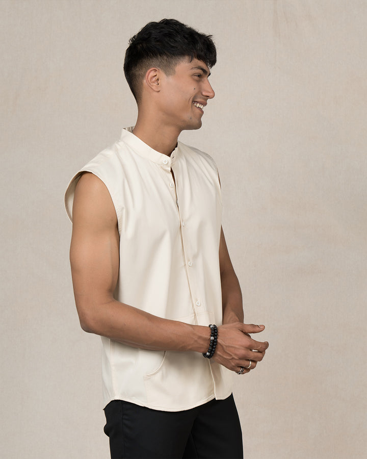 A man with short dark hair stands against a plain white background. He is smiling and wearing a Creme Brulee - Sleeveless Shirt made of 100% premium cotton and black pants. He has a black bracelet on his right wrist and is holding it with his left hand near his neck, embodying timeless style.