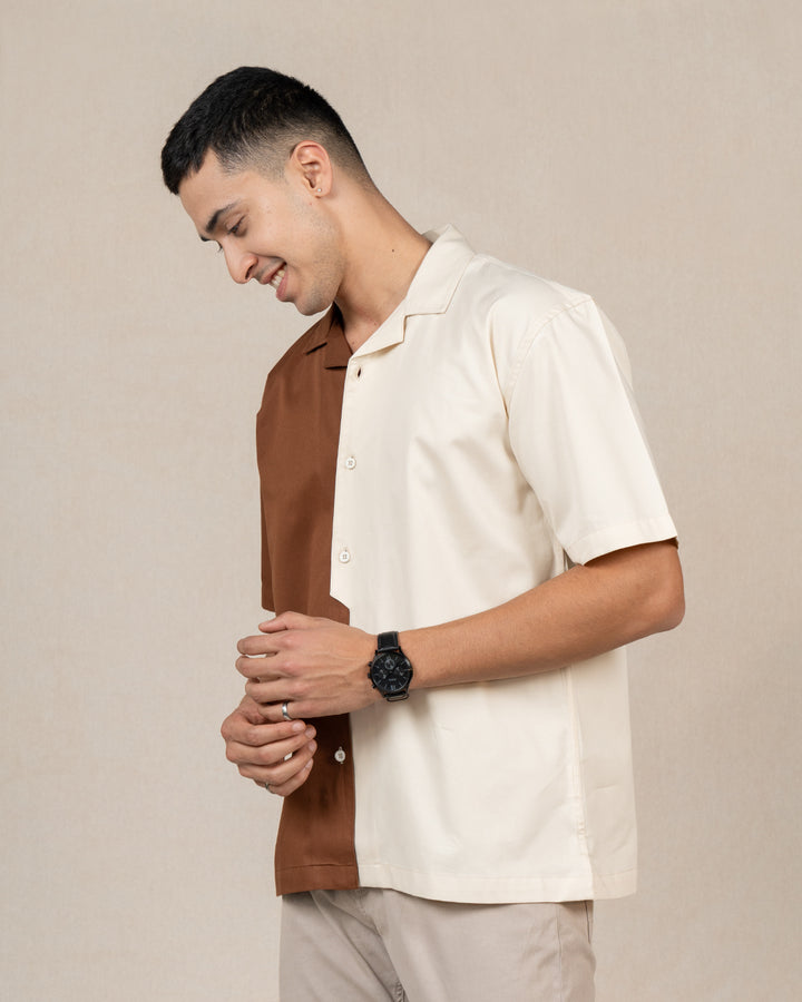 A smiling man wearing a short-sleeved Brunn Moitie - Cuban Collar Shirt from the High IQ Dye Collection. The unique design features a vertical split, with one half in rich brown and the other in light cream, made from premium cotton. He stands against a neutral background with his hands behind his back.