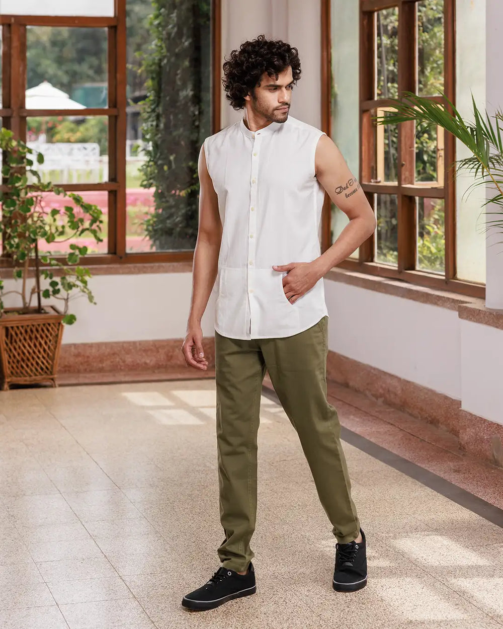 A man with curly hair is standing indoors, wearing a Branco - Sleeveless Shirt made of premium cotton and green pants. He has a tattoo on his left arm and is smiling slightly. Behind him, there are large windows with lush greenery visible outside.