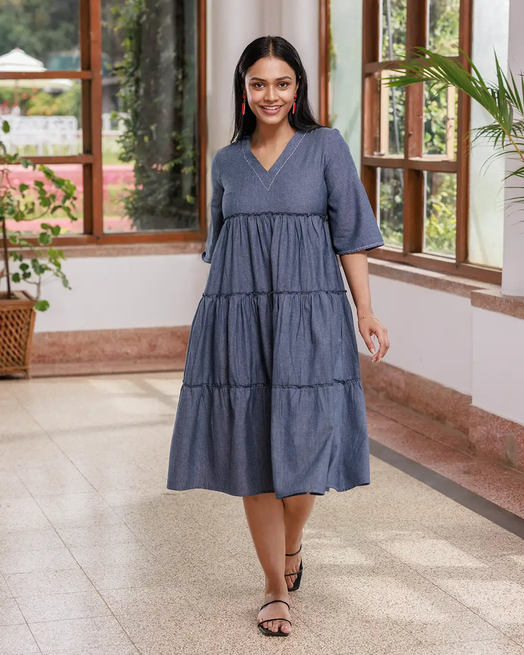 A woman with long dark hair walks inside a sunlit room. She is wearing an Opus - V Neck Flared Sleeve Dress and black strappy sandals. The room has large windows, a few green plants, and a polished floor. She is smiling confidently.