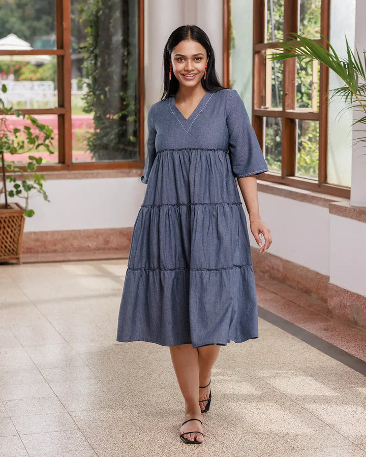 A woman with long dark hair walks inside a sunlit room. She is wearing an Opus - V Neck Flared Sleeve Dress and black strappy sandals. The room has large windows, a few green plants, and a polished floor. She is smiling confidently.
