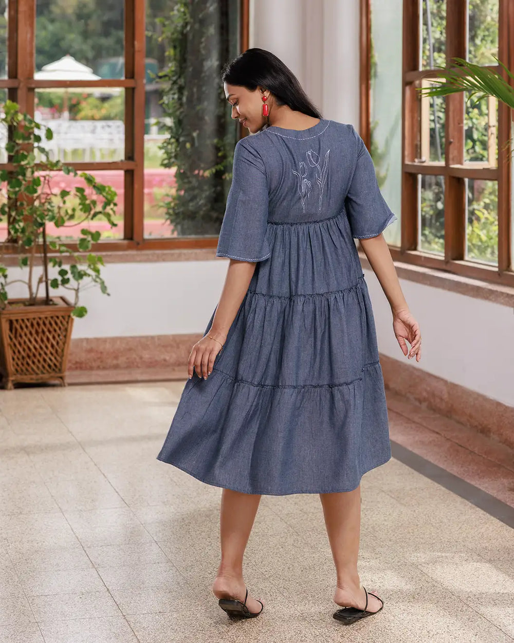 A woman with long dark hair walks inside a sunlit room. She is wearing an Opus - V Neck Flared Sleeve Dress and black strappy sandals. The room has large windows, a few green plants, and a polished floor. She is smiling confidently.