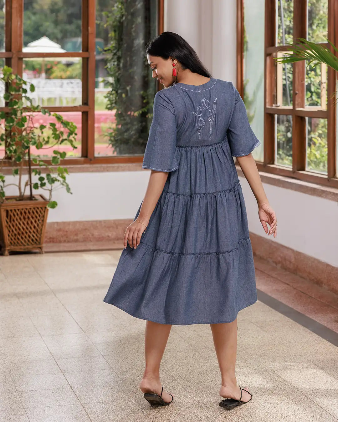 A woman with long dark hair walks inside a sunlit room. She is wearing an Opus - V Neck Flared Sleeve Dress and black strappy sandals. The room has large windows, a few green plants, and a polished floor. She is smiling confidently.