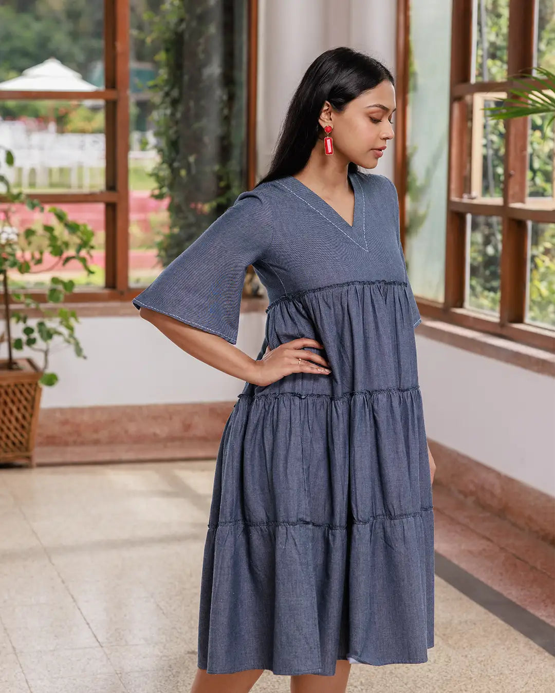 A woman with long dark hair walks inside a sunlit room. She is wearing an Opus - V Neck Flared Sleeve Dress and black strappy sandals. The room has large windows, a few green plants, and a polished floor. She is smiling confidently.